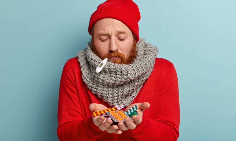 Photo of sick bearded man looks ill and exhausted, has high temperature, caught cold during winter weather, holds many pills, takes antibiotics and medicaments. Treatment and sickness concept